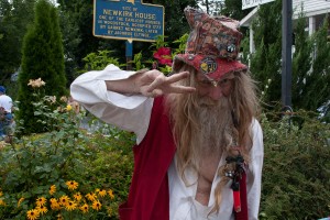 very long haired bearded man with decorated brimmed hat gives the peace sign with a shy smile