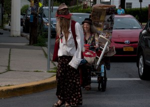 Bearded man in decorated and long skirt guides the extravagantly decorated tricycle of bearded woman and little white dog