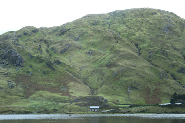 View accross the lake intense, immense green rounded mountain very small stone house at base