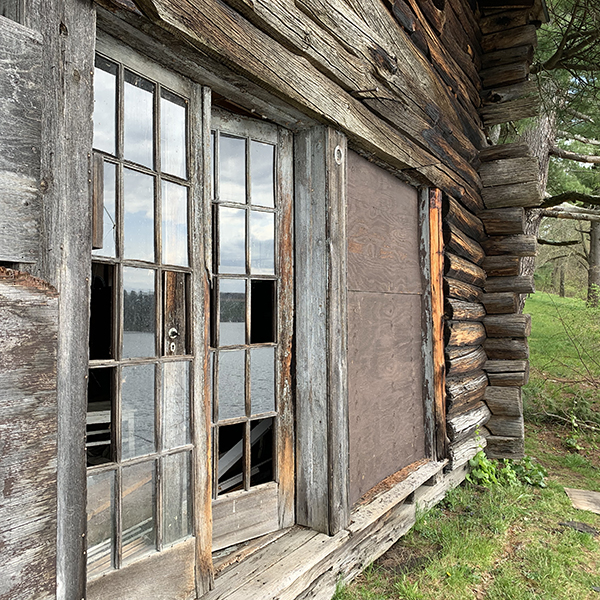 side angle of log cabin with french doors in disrepair, lake reflection in glass