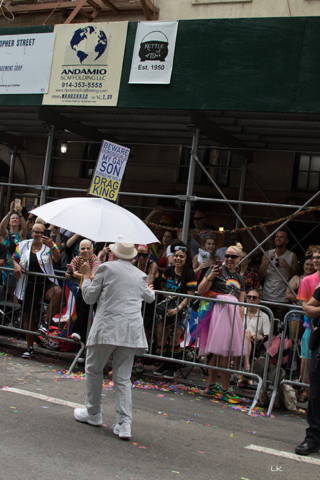 woman in man's white suit, shoes and hat holding white umbrella with sign Beware my gay son made me a drag king