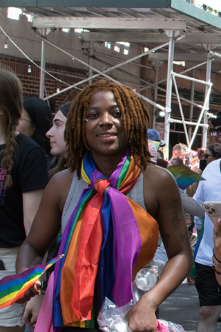 dark skinned person with reddish dreads with large scarf in pride colors around neck