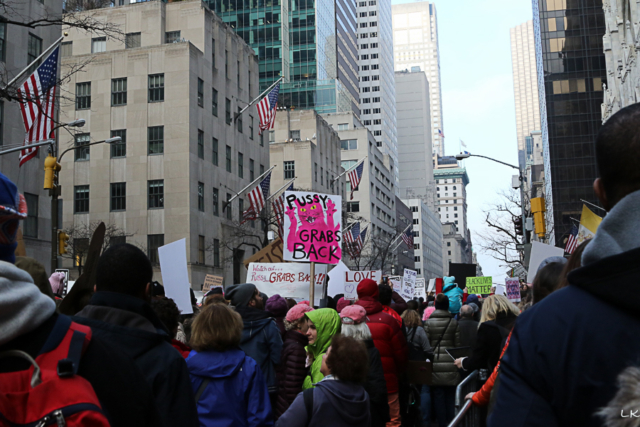 large crowd walking down NY city avenue signs held above heads one reads pussy grabs back