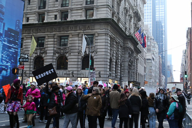 crowd of people fills intersection NYC buildings loom in background person in frong holds sign respect