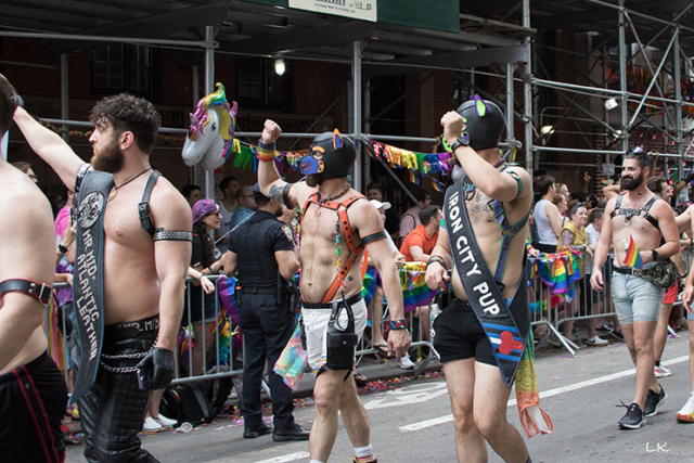 bare chested marchers wearing harnesses some with black pup masks and sign Iron City Pup