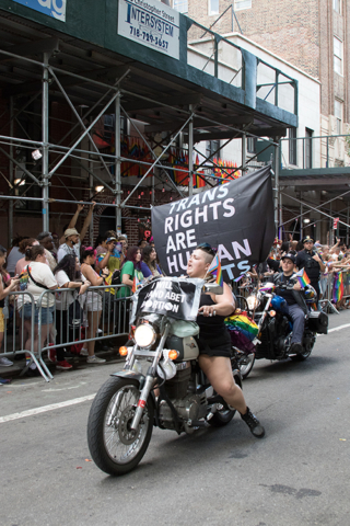 person with short blue hair  riding motorcycle with banner trans rights are human rights