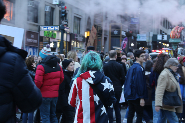 person with long green hair and wearing american flag coat shot from back among a crowd of people