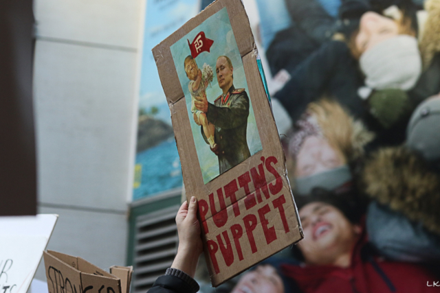 crowd with hand upraised holding cardboard sign Putins Pupped and pic of Putin in uniform holding up baby trump