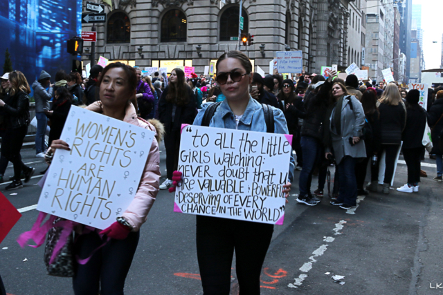 Two woman standing out from crowd holding signs womens rights are human rights and to all the little girls watching never doubt that you are valuable & powerful and deserving of every chance in the world
