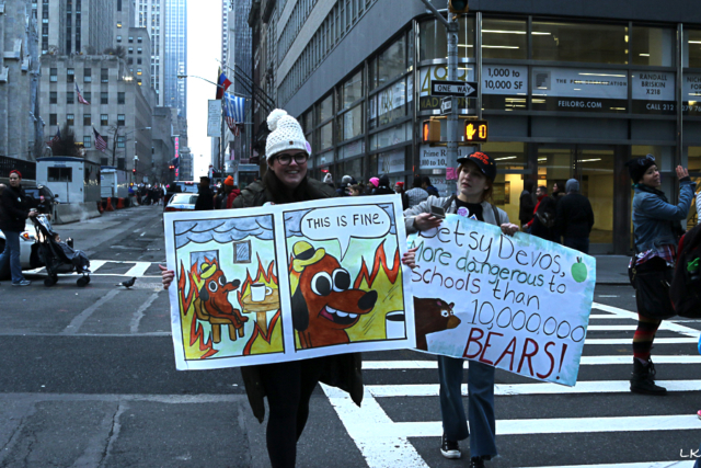 two women crossing street one holds large sign betsy devos more dangerous to schools than 10,000,000 bears