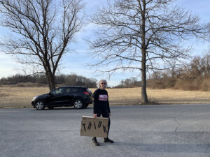 woman standing on country road holding cardboard suitcase that reads THINK