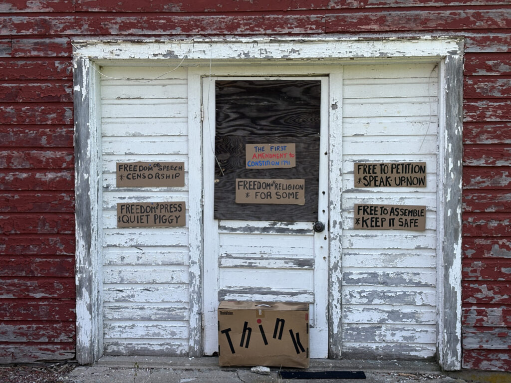 hand written cardboard signs with comments on attacks of the first amendment to constitution posted old red barn with white door, paint peeling 