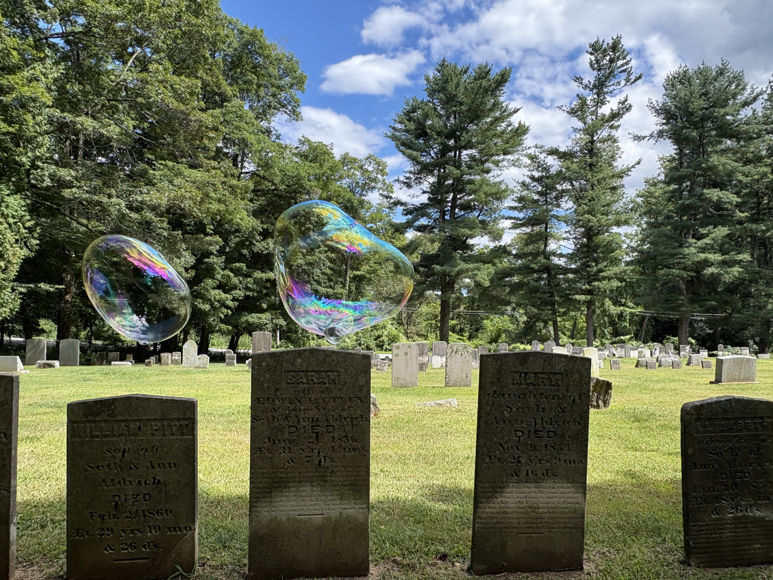 two iridescent orbs hover Sarah and William's tombstones in old cemetery background has  blue skies and large trees 